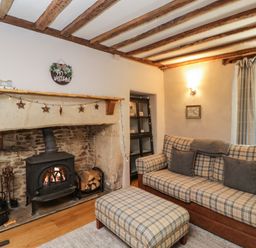 A living room with a wood stove fireplace and plaid sofa and ottoman at Fox Den Cottage in Castle Combe