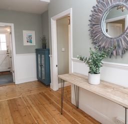 A hallway with a wooden floor a wooden console table with a plant a round sunburst mirror a blue cabinet and a white door at Belmont in Newlyn