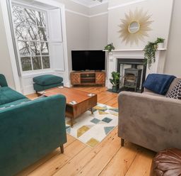 A living room with green and gray sofas a wooden coffee table a television on a wooden stand a fireplace with plants and a sunburst mirror above it at Belmont in Newlyn