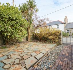 A stone paved patio area with shrubs and a palm tree near a gated entrance and stone wall at Min Y Mor - Morfa Nefyn