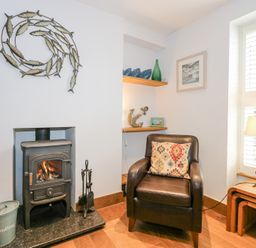 A corner with a wood burning stove a leather armchair with a patterned cushion beside a window with shutters and a small wooden lamp table at Min Y Mor - Morfa Nefyn
