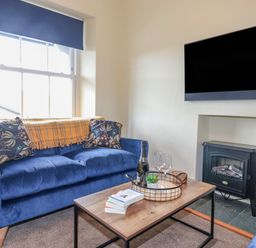 A living room with a blue sofa two patterned cushions a wooden coffee table with a tray and books a television mounted above a fireplace at Primrose Cottage in Ambleside