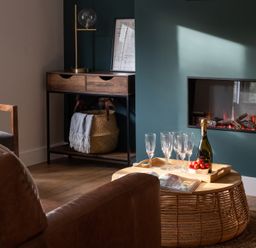 A living room with a leather chair a wooden cabinet a basket a fireplace and a wicker coffee table with champagne glasses strawberries and an open magazine at Hebog in Beddgelert