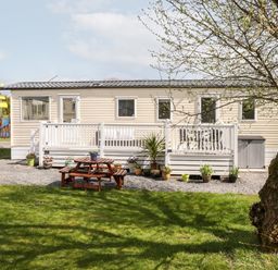 A beige mobile home with a white railing and outdoor furniture surrounded by potted plants and grass at The Maples Steel Green near Haverigg