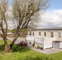 A beige mobile home with white railing and potted plants beside a tree and picnic table at The Maples in Steel Green near Haverigg