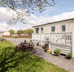 An outdoor area with a white deck attached to a mobile home and wooden picnic table on grass at The Maples in Steel Green near Haverigg