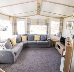 A living room with a grey sectional sofa yellow and grey cushions a television on a wooden stand and striped curtains at The Maples in Steel Green near Haverigg