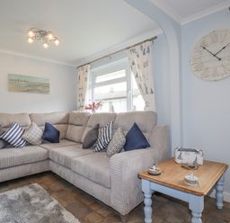A living room with a beige sectional sofa with cushions a wooden side table with decor under a large wall clock at Merryfield Lodge Rosecraddoc near Liskeard