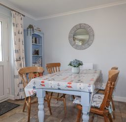 A dining area with a table covered with a lighthouse patterned cloth and wooden chairs around it with a round mirror on the wall and a blue bookshelf in the corner at Merryfield Lodge Rosecraddoc near Liskeard