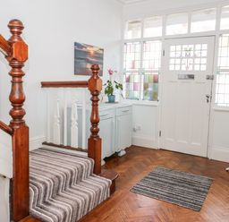 An entryway with a wooden staircase, a striped carpet runner, a small cabinet with a plant, stained glass windows, and a coat rack at 3 St Davids Place in Llandudno