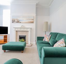 A living room with a green sofa and ottoman a fireplace a wooden TV stand and a floor lamp at 24 Cadnant Park in Conwy