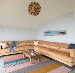 A living room with a large beige sectional sofa a wooden coffee table a colorful striped rug and a seascape painting on the wall at The Old Coastguard Lookout in Port Isaac