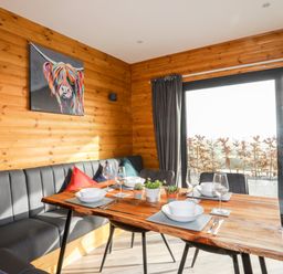 A dining area with a wooden table set with bowls wine glasses and cutlery beside a bench and chairs with cushions at Hilltop Lodge in Corwen