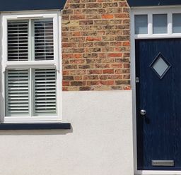 A brick wall with a white window with shutters and a dark blue door with a diamond shaped window at No 37 in Beverley