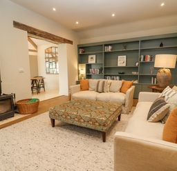 A living room with two sofas a patterned ottoman a wood burning stove and a built in bookshelf at Hawthorn Barn in Northleach