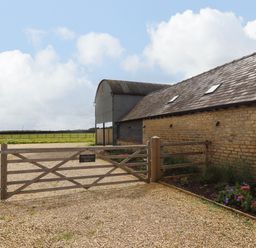 A gravel driveway with a wooden gate and a stone barn next to a flower bed at Hawthorn Barn in Northleach