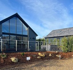 Two modern dark buildings with large glass windows and chimneys surrounded by mulch and shrubs at Foxes Den in Allerston near Thornton Dale