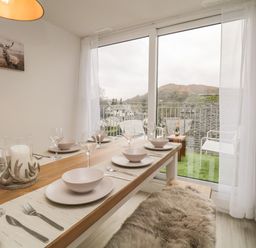 A dining area with a wooden table set with plates bowls glasses and cutlery next to a window overlooking a balcony and hills at Dixon Court in Ambleside