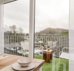 A dining table set with plates and glasses next to a balcony with chairs and a small wooden table overlooking houses and hills at Dixon Court in Ambleside