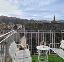 A balcony with two white chairs and a small table holding a bottle and glasses overlooking rooftops and hills at Dixon Court in Ambleside