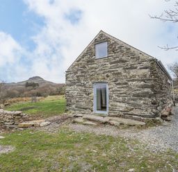 A stone building with two windows in a rural area with grass trees and a hill in the background at Hen Beudy in Porthmadog