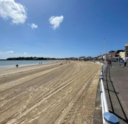 A sandy beach with tire tracks and footprints beside a paved promenade with pedestrians and buildings at Sunrise Apartment in Weymouth