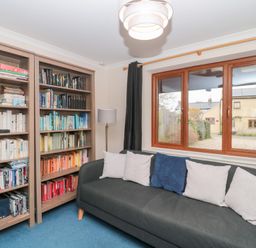 A room with bookshelves filled with books and DVDs a gray sofa with white and blue cushions a window with a view of houses and a lamp next to the sofa at Morgan House in Middlezoy