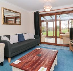 A living room with a dark sofa and cushions a wooden coffee table a TV on a wooden stand and glass sliding doors leading to a conservatory at Morgan House in Middlezoy