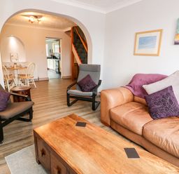 A living room with a brown leather sofa and two gray chairs around a wooden coffee table with a dining area and kitchen visible through an arch at 57 Southend Road Hunstanton