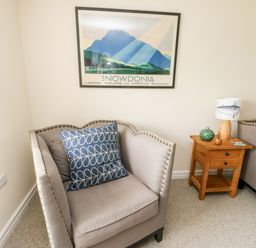 A corner chair with cushions next to a wooden side table with a lamp and decoration under a snowdonia framed picture at 8 Llwyn Onn in Llanfairpwllgwyngyll
