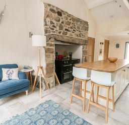 A kitchen with a stone fireplace a blue armchair a wooden island with two stools and a patterned rug at 8 Llwyn Onn in Llanfairpwllgwyngyll