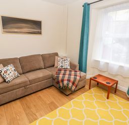 A living room with a brown sectional sofa with pillows and a blanket a wooden small table yellow patterned rug and blue curtains at Ty Cwtch in Tenby