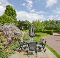 An outdoor patio with a glass table and six chairs with a closed umbrella near a brick wall with flowers and a garden at The Stables in Belbroughton