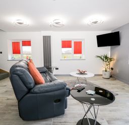 A living room with a dark leather sofa and orange cushions two round tables a wall mounted TV windows with red blinds and a potted plant at The Annex in Ulverston