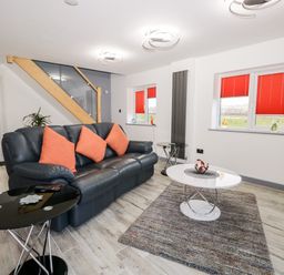 A living room with a black leather sofa and orange cushions with two round tables a rug and red window blinds at The Annex in Ulverston