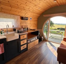 A small kitchen and living area with wooden walls and ceiling a sofa and round glass door opening to a patio at Dew Pond in Blagdon