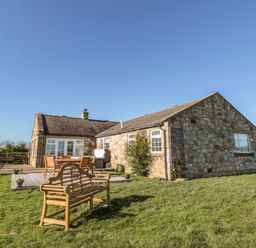 A stone house with a wooden bench and outdoor dining set on a lawn at Thistleyhaugh Cottage in Longhorsley near Longframlington