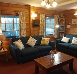 A living room with two blue sofas and floral cushions a wooden coffee table with flowers a wooden side table with a lamp and floral curtains at Orchard Lodge in Hockworthy near Sampford Peverell Devon