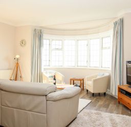 A living room with a beige sofa two white armchairs a wooden TV stand with a television tripod floor lamp wall clock and large windows with curtains at Ocean Watch 5 in Greenhill