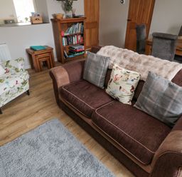 A living room with a brown sofa and patterned armchair a wooden bookshelf and dining table at Castle Keep in Norham