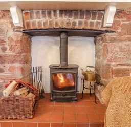 A wood burning stove with fire inside a stone fireplace with a basket of firewood and fireplace tools nearby at Rose Cottage in Holcombe in Teignmouth