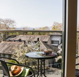 A balcony with a round table holding a bottle, two glasses, a hat, and sunglasses overlooking rooftops and trees at Claife Heights in Windermere