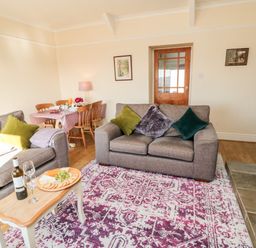 A living room with a patterned rug gray sofas green cushions a wooden coffee table with wine and cheese and a dining table with chairs at Healey Farm Cottage in Rothbury