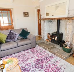 A living room with a grey sofa and cushions a fireplace with a stove and a wooden mantle a rug on the floor and a wooden table with wine and snacks at Healey Farm Cottage in Rothbury