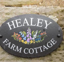 A black oval sign with white text Healey Farm Cottage and painted flowers on a stone wall at Healey Farm Cottage in Rothbury