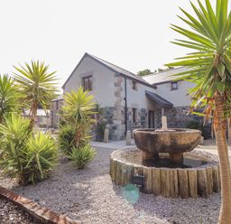 An outdoor garden with palm-like plants gravel ground and a stone water fountain in front of a stone and white plaster house at Polurrian House in Cury near Mullion