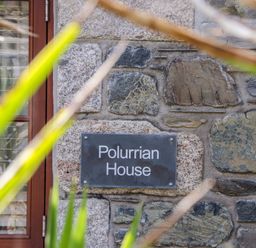 A stone wall with a sign that says Polurrian House next to a wooden door partially obscured by green plants at Polurrian House in Cury near Mullion
