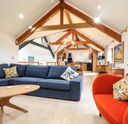 A living area with a blue sectional sofa, an orange armchair, and a wooden coffee table under wooden ceiling beams at Polurrian House in Cury near Mullion