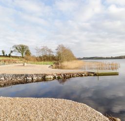 A lakeside area with a pebble beach and a wooden dock surrounded by reed beds at The Water's Edge in Boyle County Roscommon