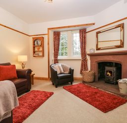 A living room with a brown leather sofa a black armchair a fireplace with a wood stove red rugs and curtains at The Lodge in Greystoke